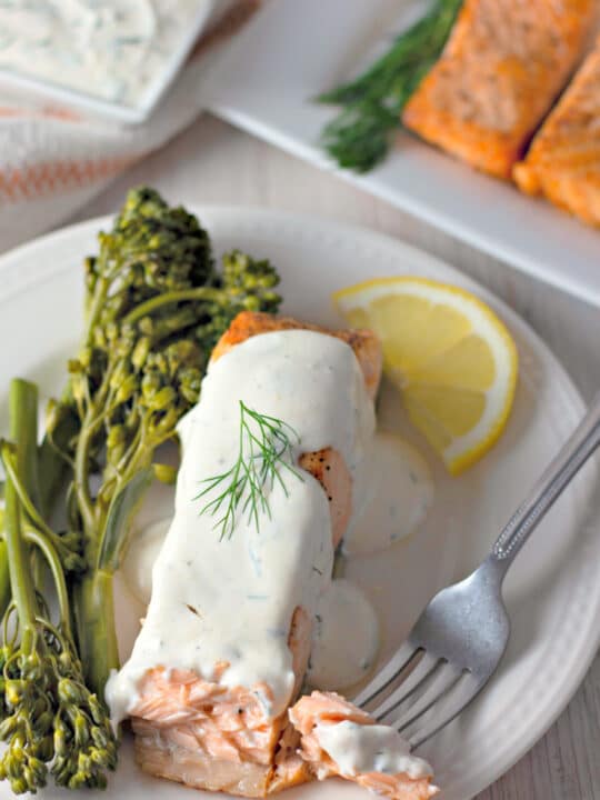 salmon in creamy dill sauce on white plate with broccolini and lemon slice