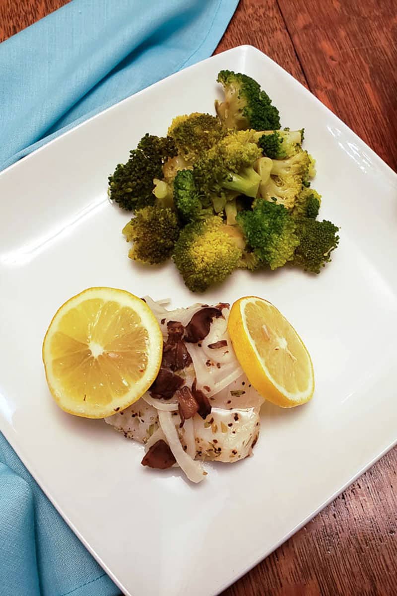 Baked tilapia with lemon slices and broccoli on white plate with blue napkin.