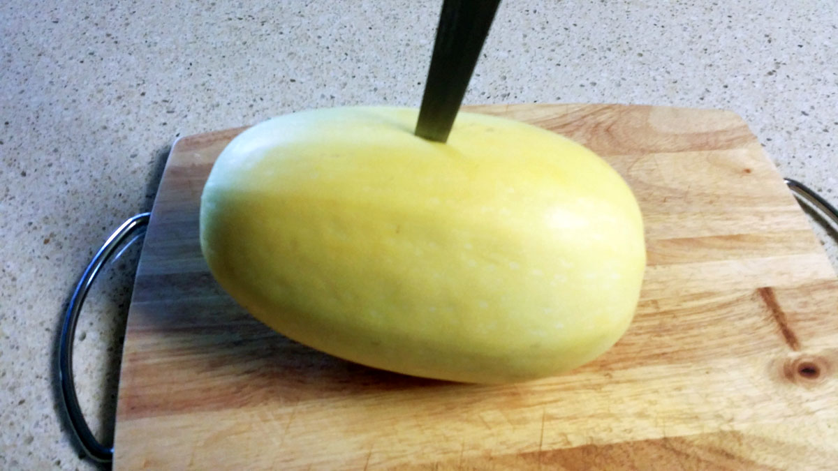 Whole spaghetti squash on cutting board with knife poking holes.